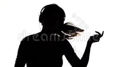 一个年轻快乐的女人戴着大耳机<strong>听</strong>音乐，拿着智能手机的<strong>剪影</strong>，女孩在白色的隔离巴上跳舞